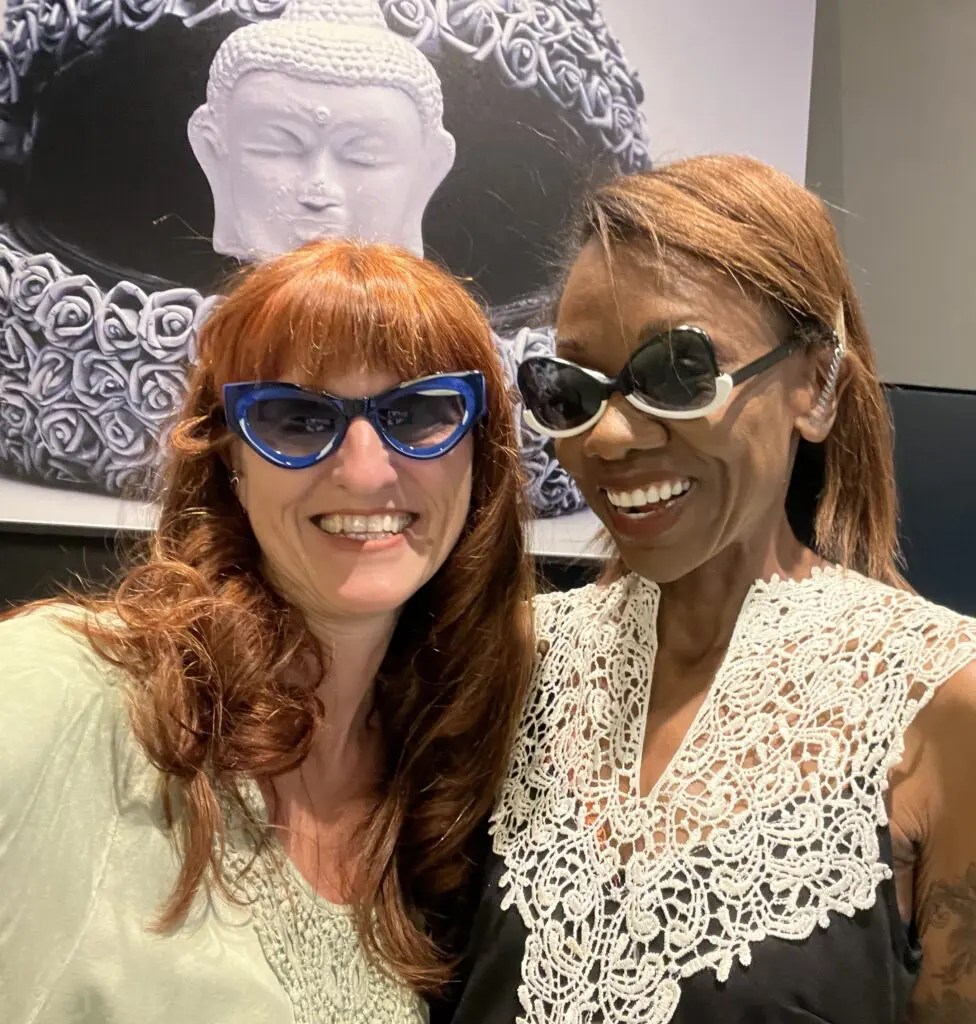 two smling women in art sunglasses in front of a painting depicting a stylized motorcycle helmet and a buddha head
