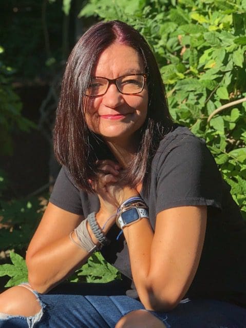 A woman with glasses sitting in front of some bushes