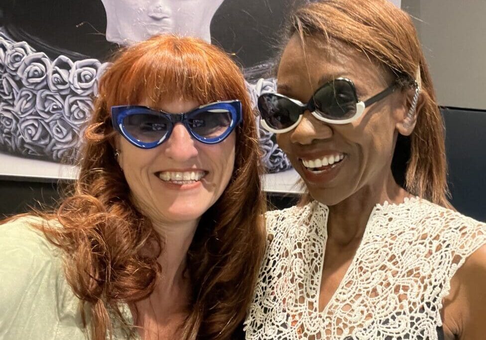 two smling women in art sunglasses in front of a painting depicting a stylized motorcycle helmet and a buddha head